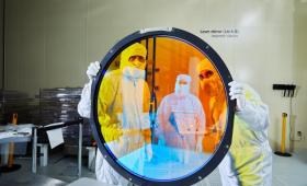 Two bunny-suited engineers look through large circular glass.