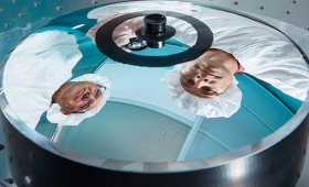 A reflection of Brian Bauman (left), the space hardware principal optical engineer and inventor of the monolithic telescope and Frank Ravizza, the space hardware optical engineering lead, is seen on the primary mirror surface on a flight-ready 175-millimeter aperture monolithic telescope. Addittionally, Ravizza is seen holding a 25-millimeter aperture monolithic optic. The ease of handling showcases the robust design incorporated in all monolithic telescopes. 