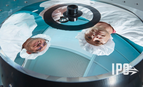 A reflection of Brian Bauman (left), the space hardware principal optical engineer and inventor of the monolithic telescope and Frank Ravizza, the space hardware optical engineering lead, is seen on the primary mirror surface on a flight-ready 175-millimeter aperture monolithic telescope. Additionally, Ravizza is seen holding a 25-millimeter aperture monolithic optic. The ease of handling showcases the robust design incorporated in all monolithic telescopes.
